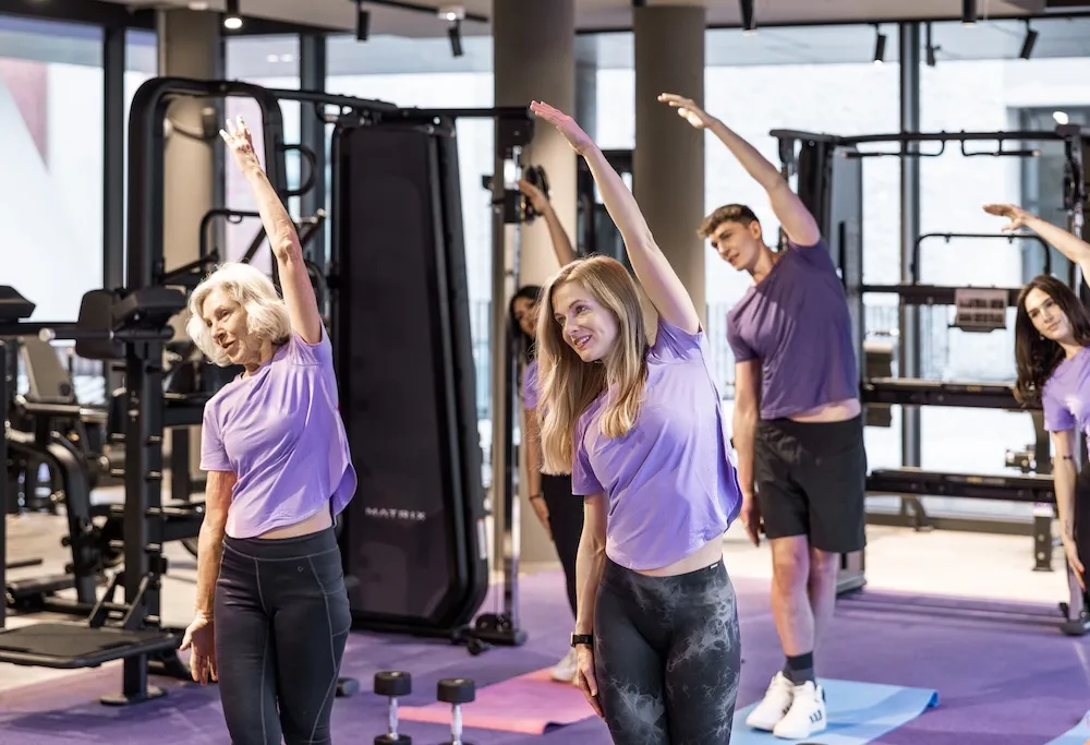Eine Gruppe von vier Personen in einem Fitnessstudio, die lila Hemden und schwarze Trainingshosen oder -shorts tragen, mit einem Arm über dem Kopf Seitenstrecken machen und auf Gymnastikmatten vor Fitnessgeräten stehen.