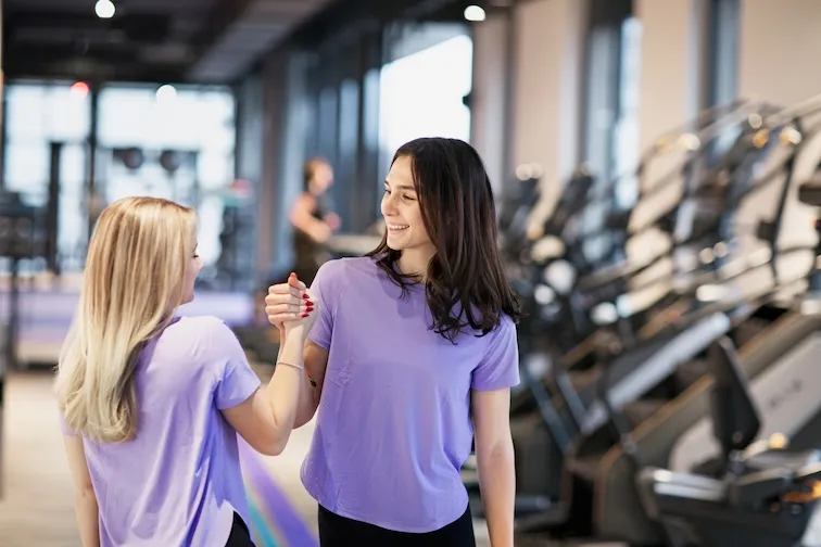 Zwei Frauen in lila Hemden lächeln und einen freundlichen Handschlag in einem Fitnessstudio mit Trainingsgeräten im Hintergrund machen.