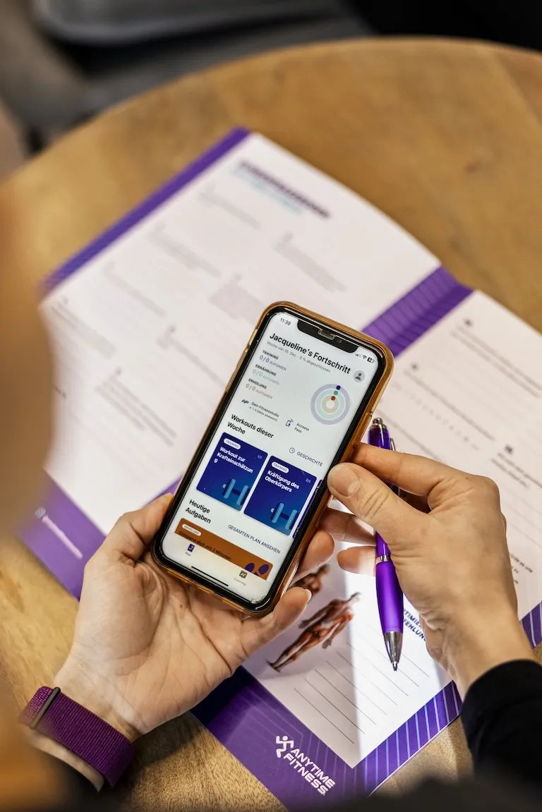Person, die ein Smartphone in der Hand hält, auf der eine Fitness-App angezeigt wird, auf der Jacquelines Fortschritt, Trainingsplan und Aufgaben für die Woche angezeigt werden, mit einem lila Stift in der anderen Hand und einem Flyer der Marke Anytime Fitness auf einem Holztisch.