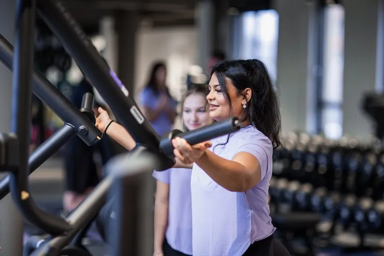 Zwei Frauen in einem Fitnessstudio; eine Frau davor benutzt eine Brustpresse, während die andere zuschaut.