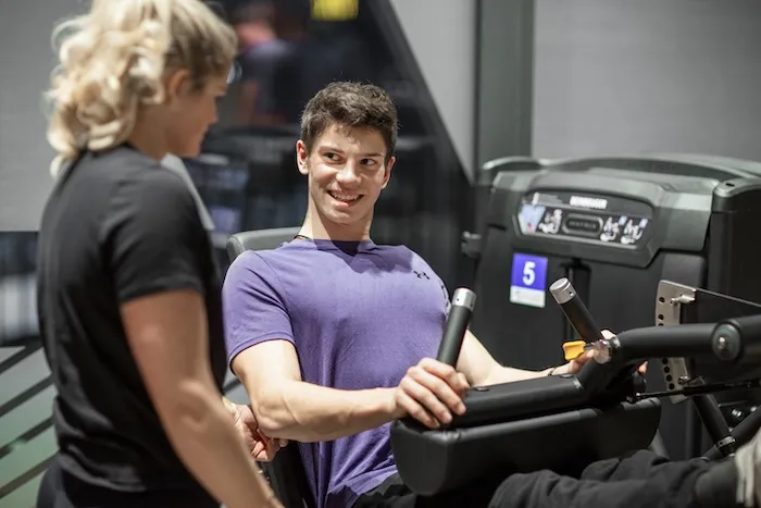Junger Mann in einem lila Hemd benutzt in einem Fitnessstudio eine sitzende Beinpresse, während er lächelt und eine Trainerin ansieht, die neben ihm steht.
