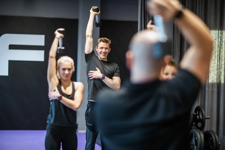 Ein Gruppenfitnesskurs, in dem ein Mann und eine Frau Hanteln mit einem Arm über den Kopf halten und dabei den anderen Arm über die Brust strecken. Sie folgen einem Instruktor, der teilweise im Vordergrund sichtbar ist und auch Hanteln hält.