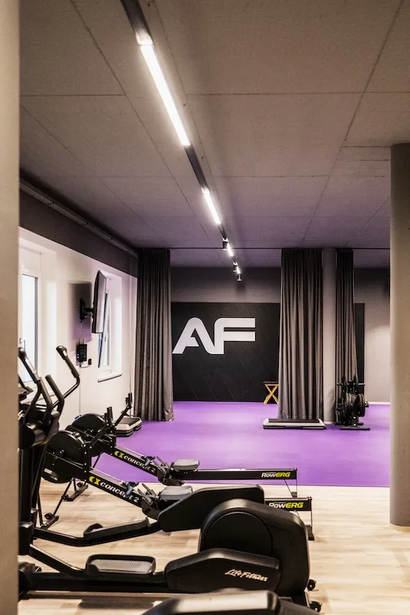 Indoor-Fitnessstudio mit zwei Concept2-Rudergeräten im Vordergrund und einem lila Trainingsbereich im Hintergrund mit einer schwarzen Wand mit großen weißen Buchstaben „AF“.