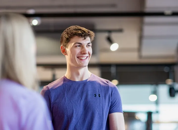 Junger Mann in einem lila Under Armour-Shirt lächelt und schaut in einem Fitnessstudio mit verschwommener Person im Vordergrund zur Seite.