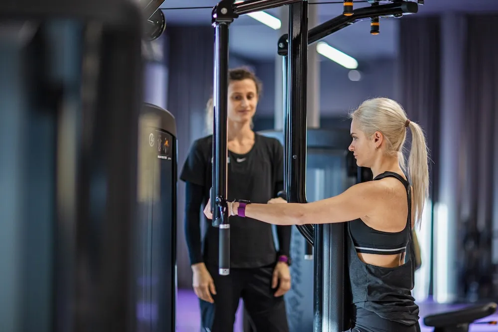 Eine blonde Frau in Sportkleidung, die eine Kabelmaschine für ein Training in einem Fitnessstudio benutzt, während ein Fit-Trainer in Schwarz ihr Training beobachtet und unterstützt.
