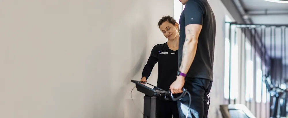 Eine Fitnesstrainerin in schwarzer Sportkleidung verwendet einen Touchscreen auf einem Laufband, während in einem Fitnessstudio mit Tageslicht ein Mann neben ihr steht und Laufbandgriffe hält.