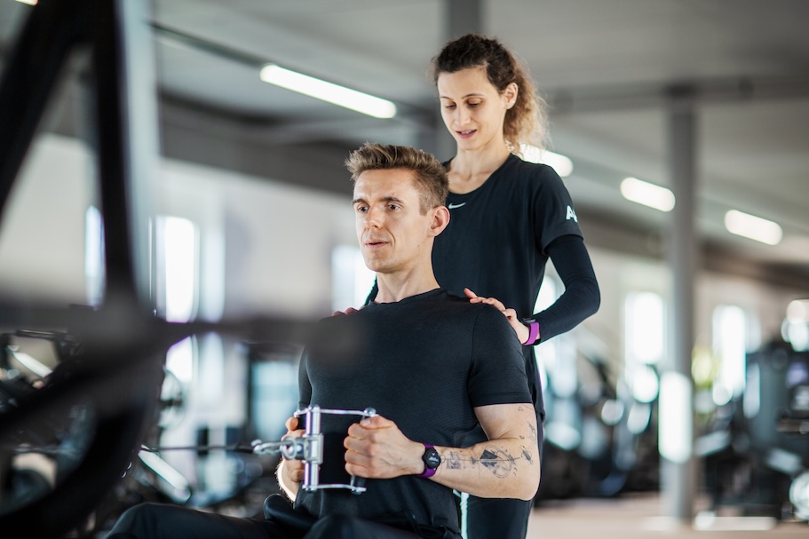 Mann trainiert an einem sitzenden Rudergerät, hinter ihm steht eine weibliche Personal Trainerin, die seine Schultern in einem geräumigen Fitnessstudio lenkt.