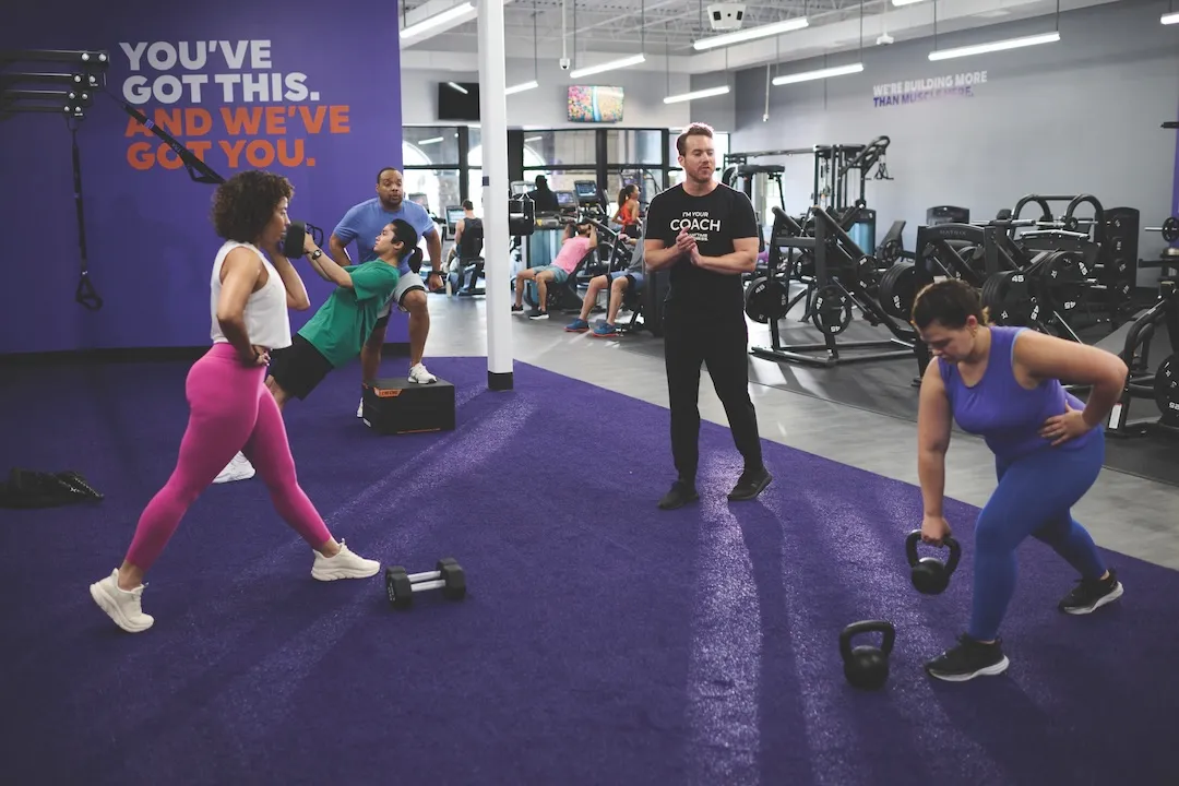 A personal trainer smiles while supervising a woman performing a dumbbell chest press on a weight bench in a modern gym.