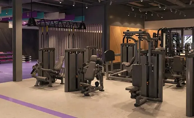 Interior de un gimnasio moderno con varias máquinas de entrenamiento de fuerza dispuestas en un piso de color claro, con una franja morada e iluminación ambiental en el techo.