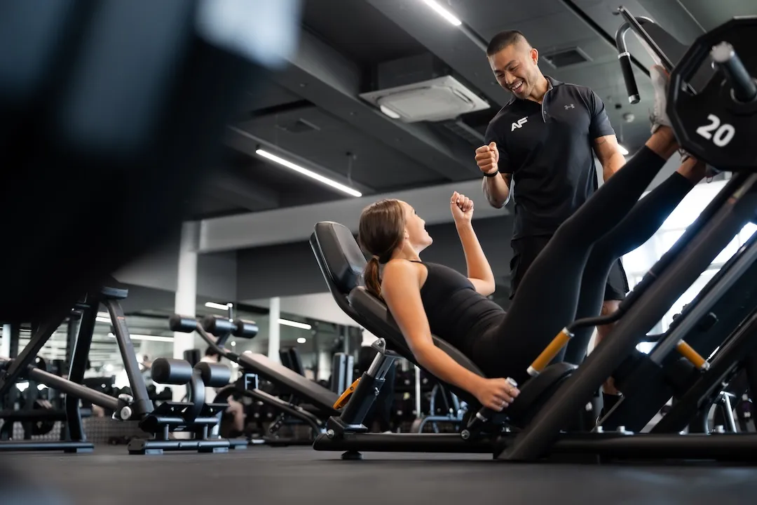 A female fitness trainee using a leg press machine while a male personal trainer in a black shirt encourages her in a modern gym.