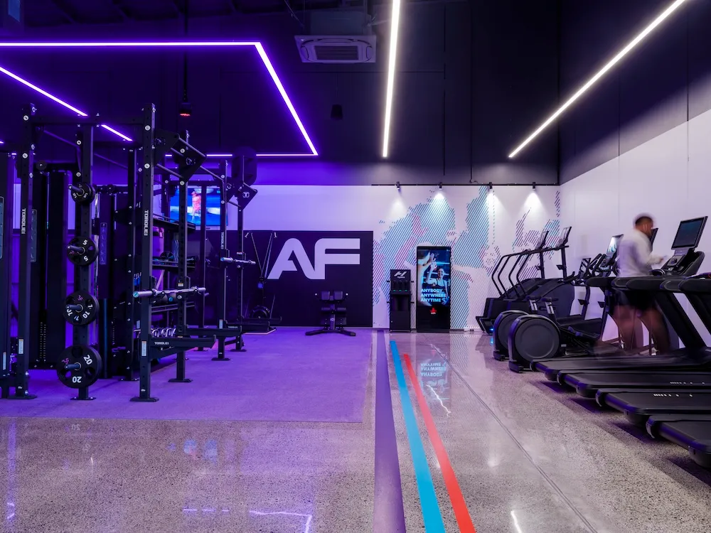 Modern gym interior with neon purple lighting, weightlifting equipment on the left, treadmills on the right with a person running on one, and a large AF logo on the back wall.