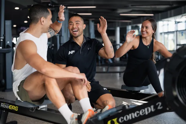 Three people in workout clothes sitting on rowing machines in a gym, smiling and giving each other high fives.