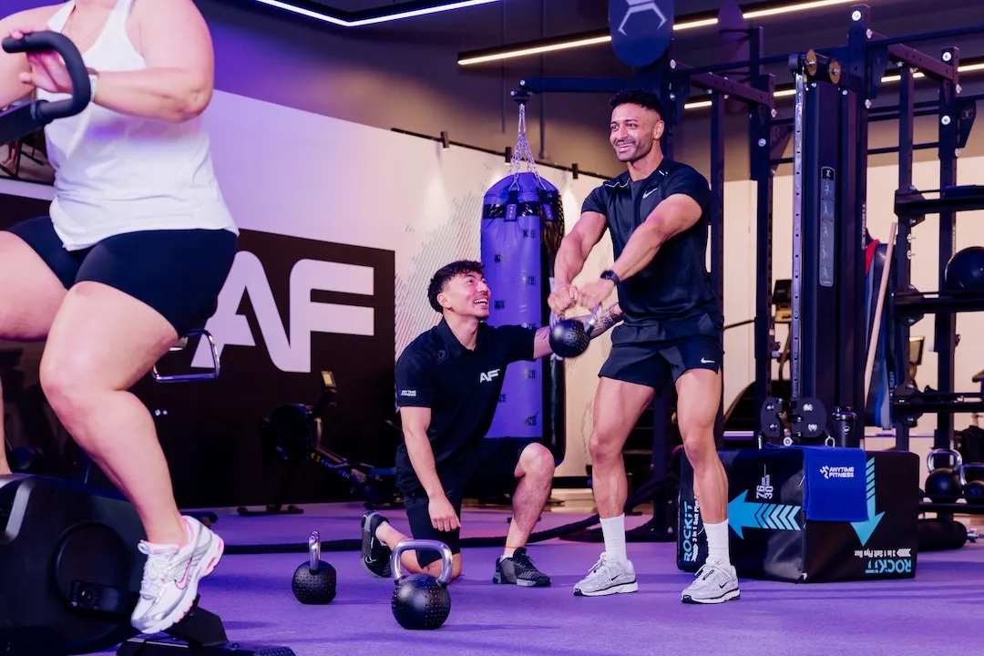 A gym scene showing a man in black athletic wear assisting another man lifting kettlebells, while a woman exercises on a stationary bike in the foreground.