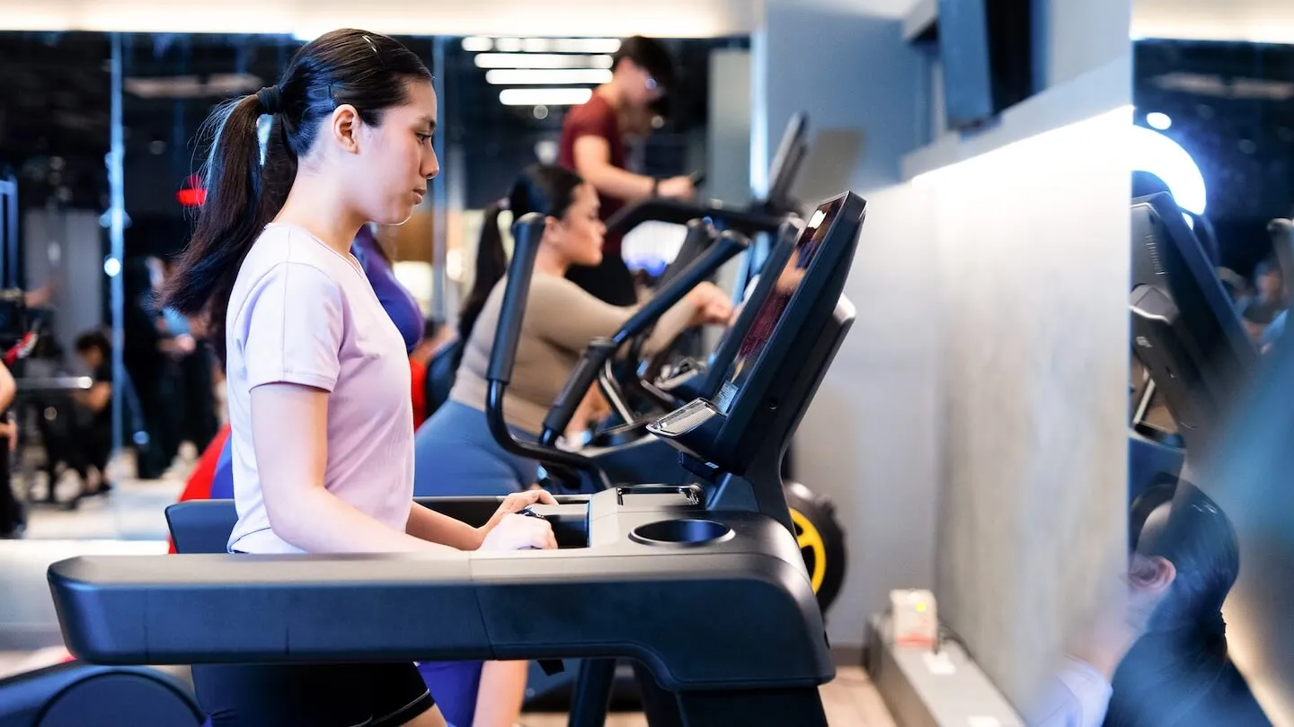 Woman using a treadmill alongside other gym members on cardio machines.