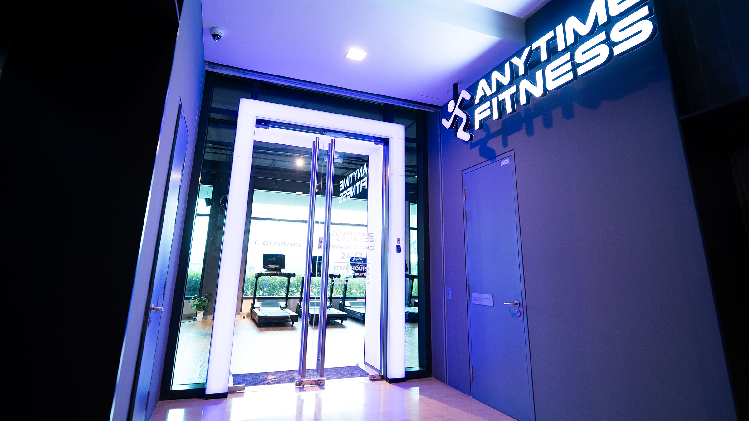 Entrance to an Anytime Fitness gym with illuminated glass doors and a bright sign above a closed door next to it; treadmills and large windows are visible inside.