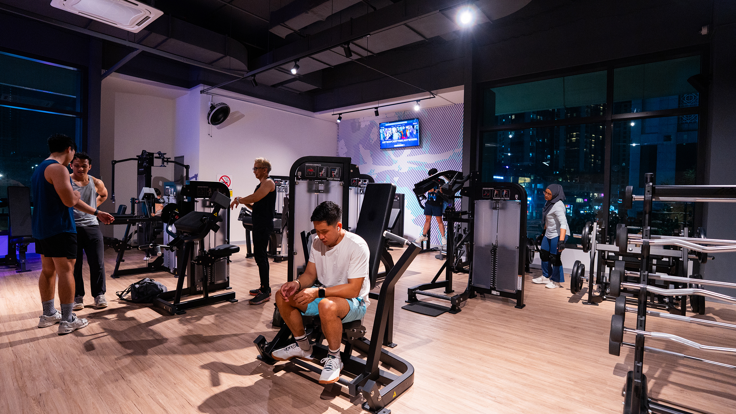 Interior of a modern gym with six people using exercise machines and equipment, including weight lifts and resistance machines, with large windows showing city buildings at night.