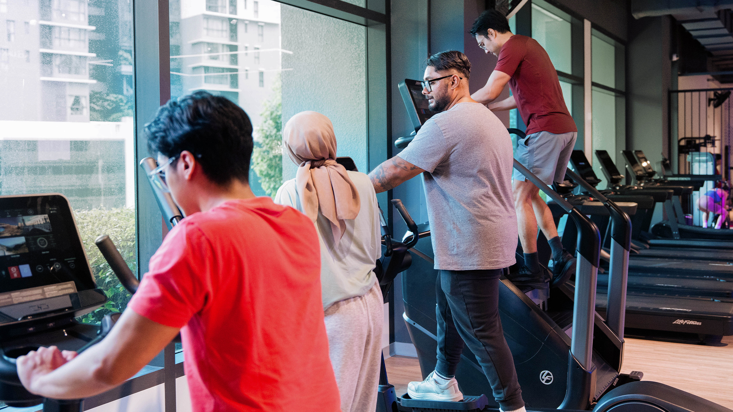 Four people exercising on cardio machines at a gym with large windows showing a city view outside; two men and a woman wearing a hijab are using different machines focused on their workouts.