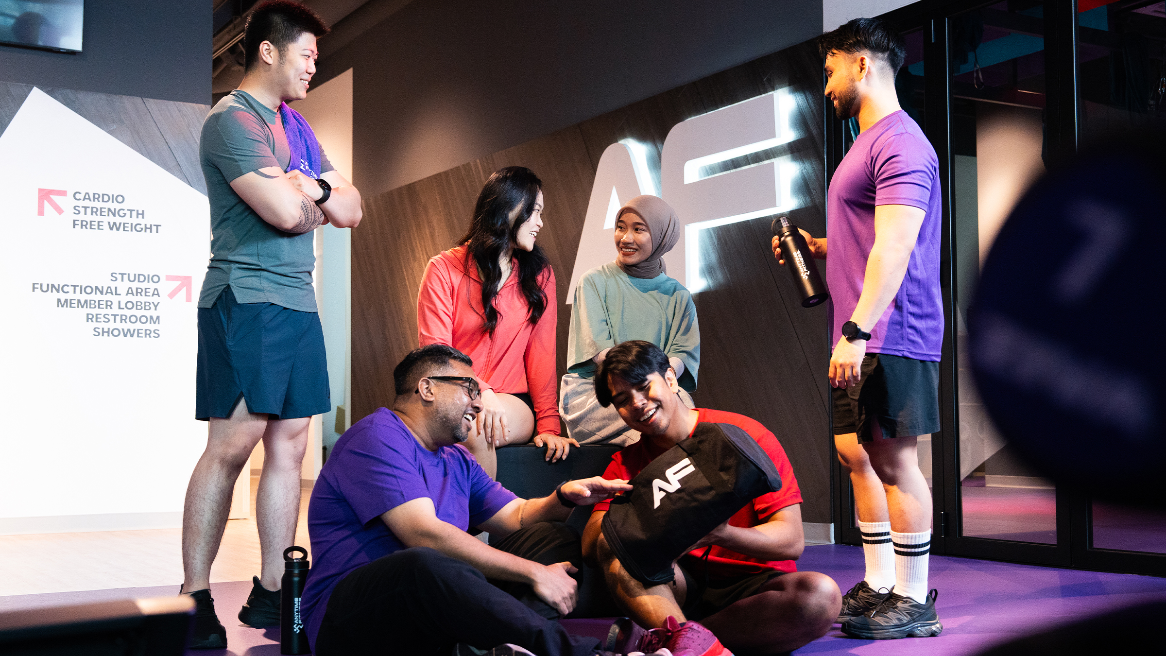 Five diverse young adults in athletic wear gather and smile in a gym setting with a large illuminated AF logo in the background, near a sign pointing to cardio, strength, free weight areas, studio, functional area, member lobby, restroom, and showers.