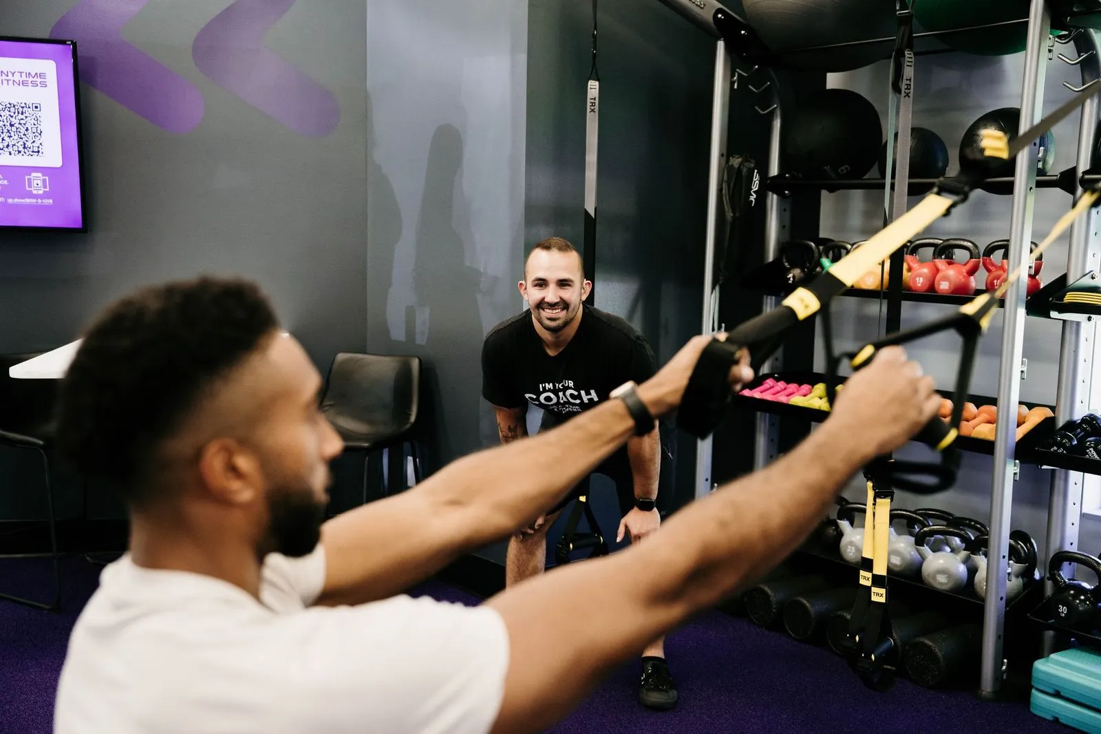 Hombre con camisa blanca que usa correas de suspensión TRX para hacer ejercicio en un gimnasio mientras un entrenador sonriente con camisa negra lo observa y lo alienta, con coloridas pesas rusas y equipos de ejercicio en el fondo.