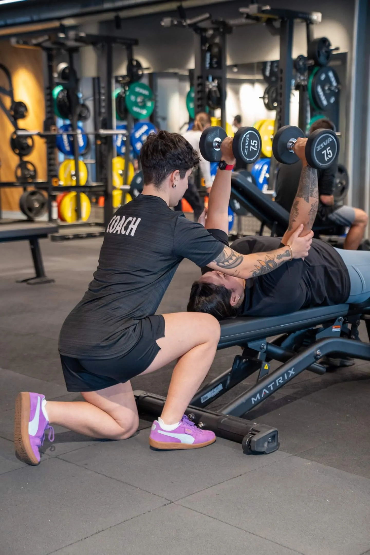Una mujer realiza un press de banca con mancuernas con la ayuda de su entrenador en el gimnasio.