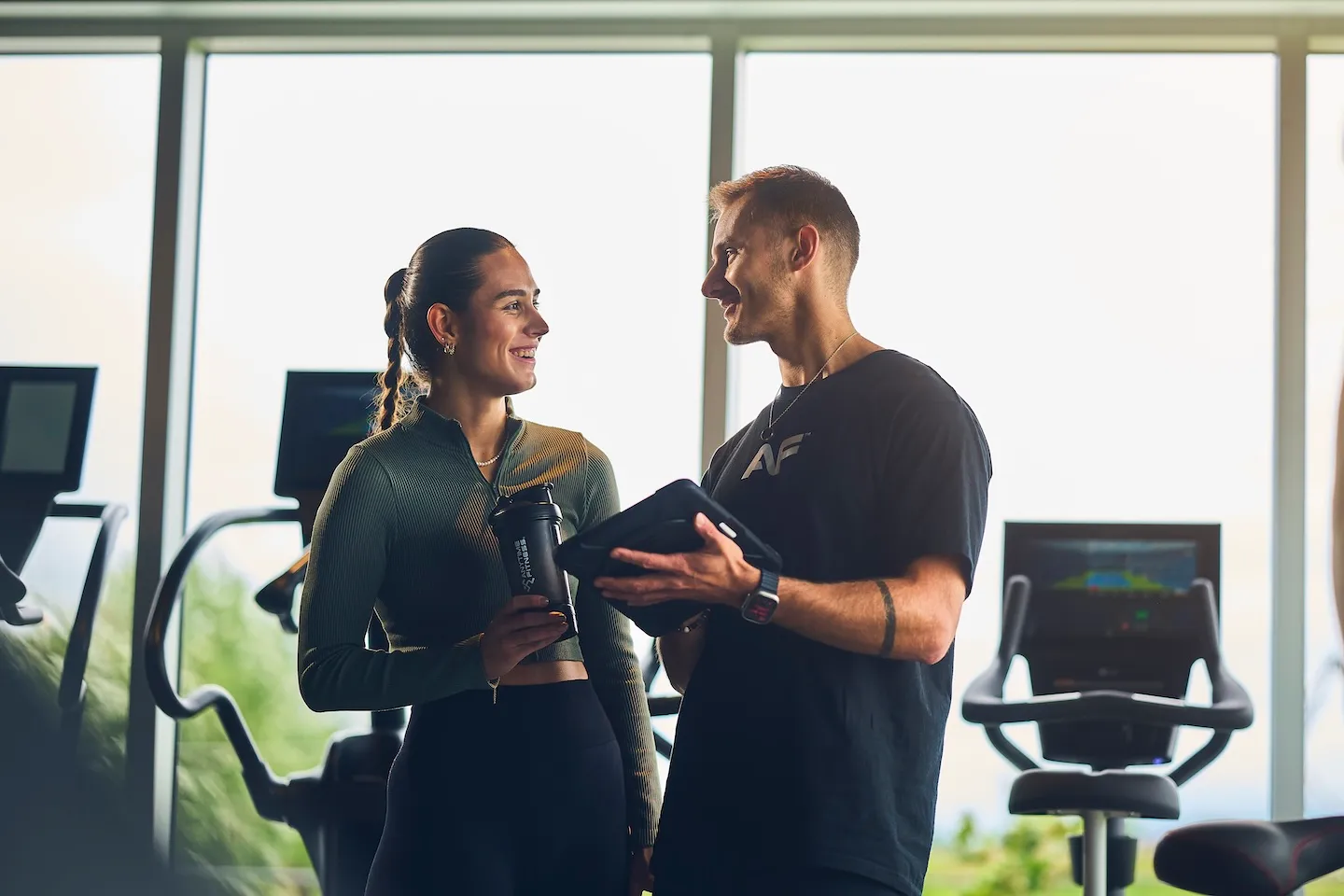 Anytime Fitness coach and member smiling together in the gym while reviewing a training plan on a tablet; the member holds an Anytime Fitness shaker bottle.