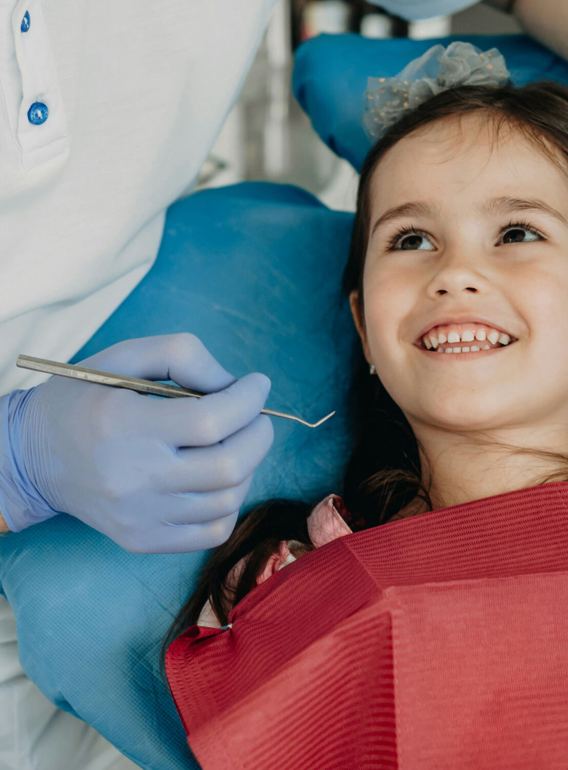 Smiling young girl reclined in dental chair with dentist's gloved hand holding dental tool beside her.