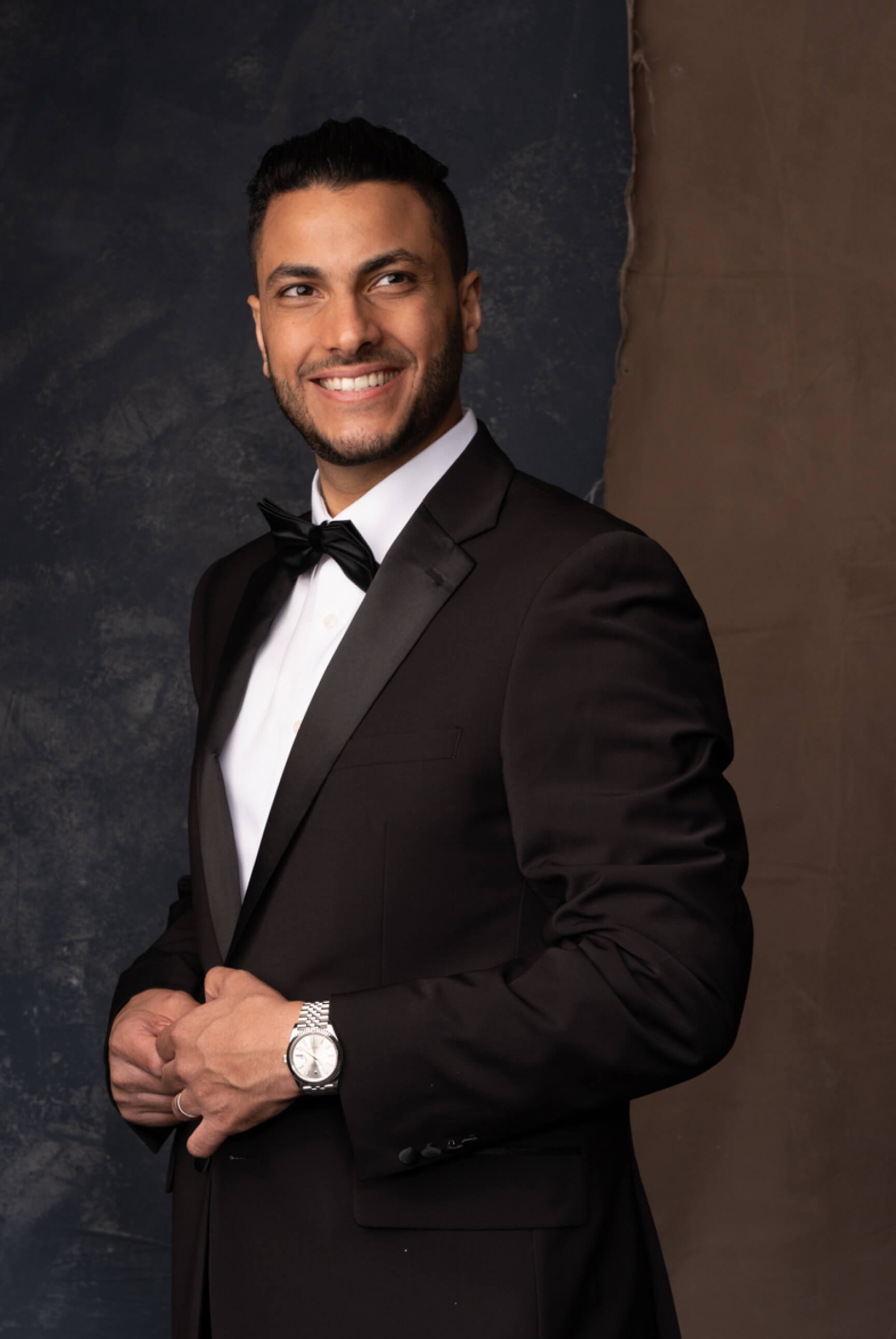 Smiling man in a black tuxedo with bow tie and silver watch standing against a two-tone background.