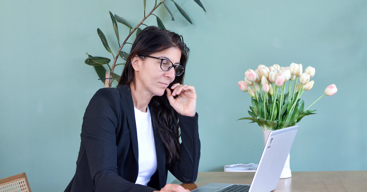 Woman at a desk with a laptop, pausing in thought