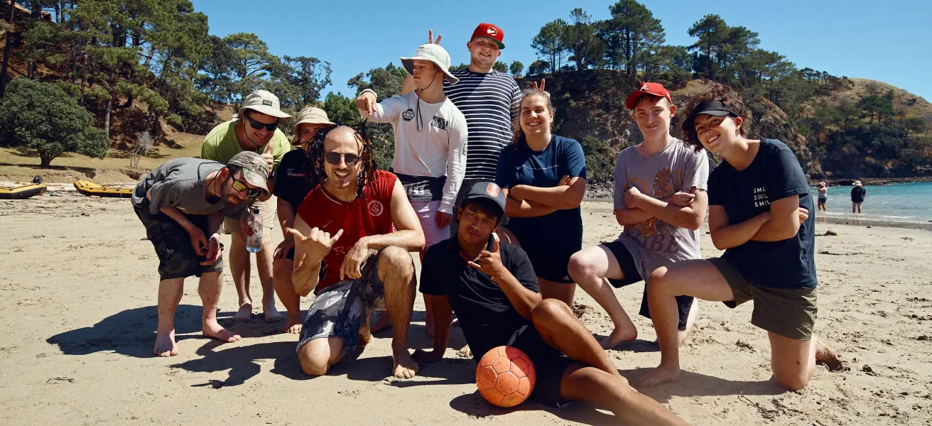 Ten participants are pictured posing for a group photo on the beach. A shoreline and trees are visible in the background.