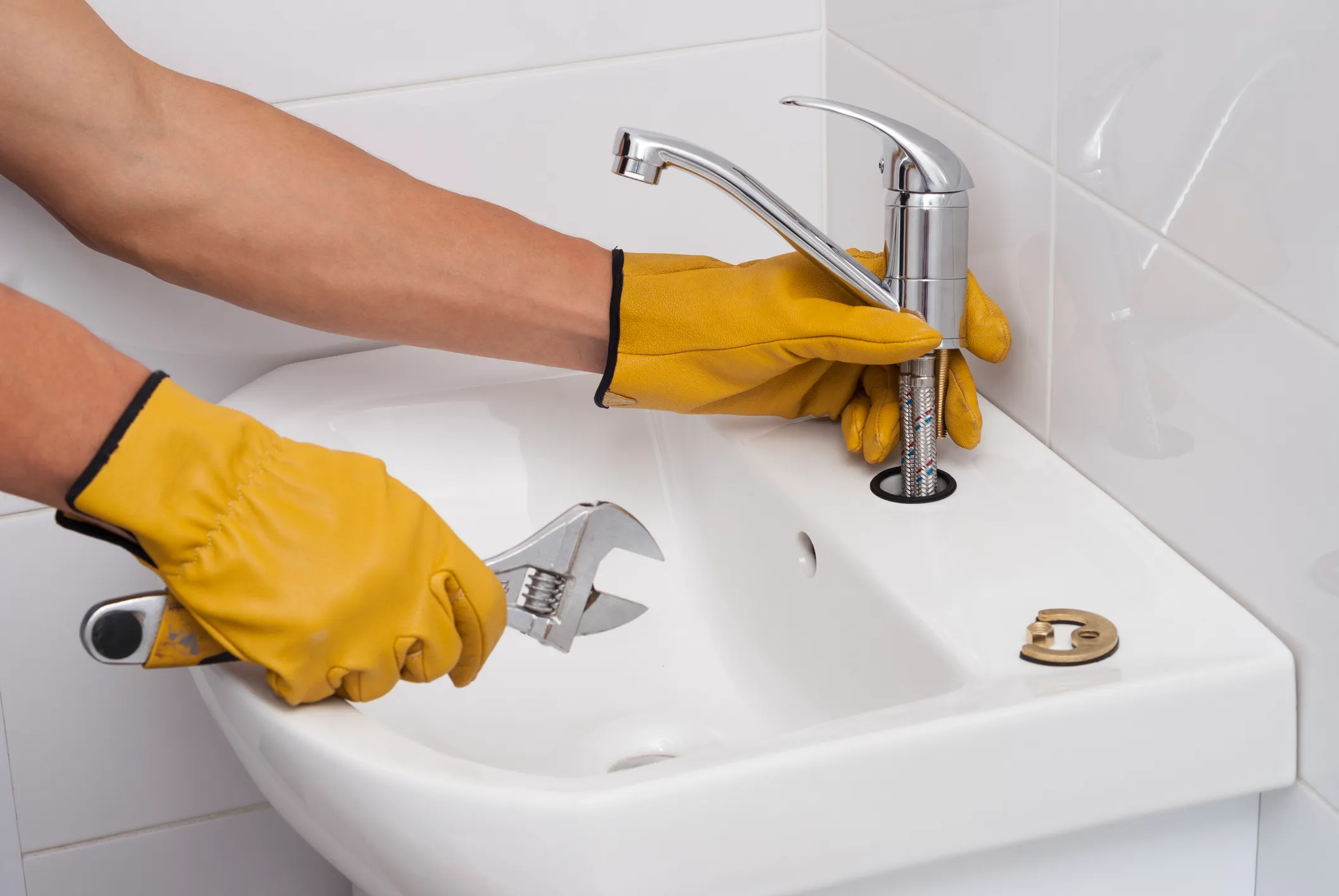 Plumber in yellow gloves installing faucet on white sink using wrench, with tiled background