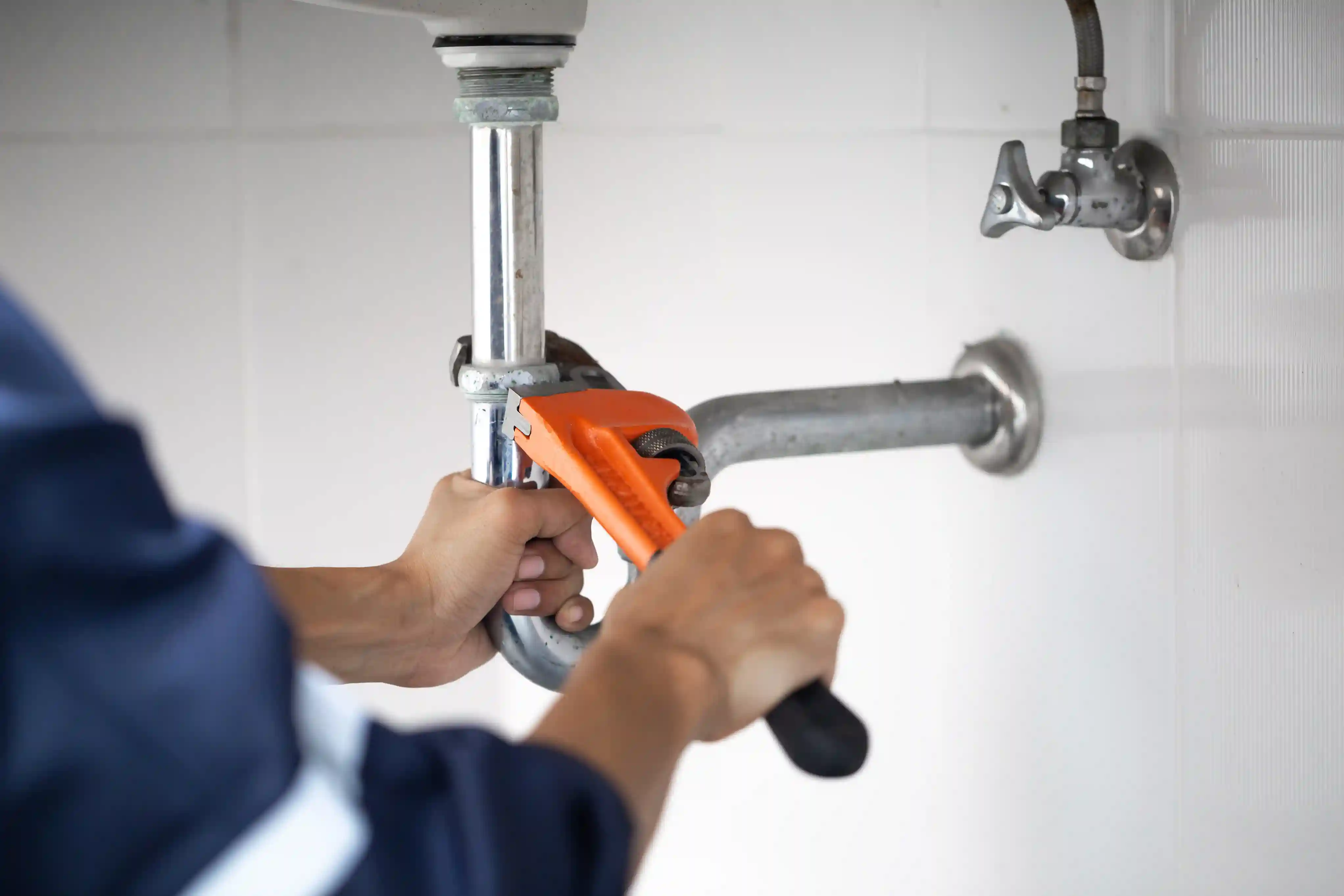 Plumber using orange pipe wrench on metal pipe under sink with wall-mounted faucet
