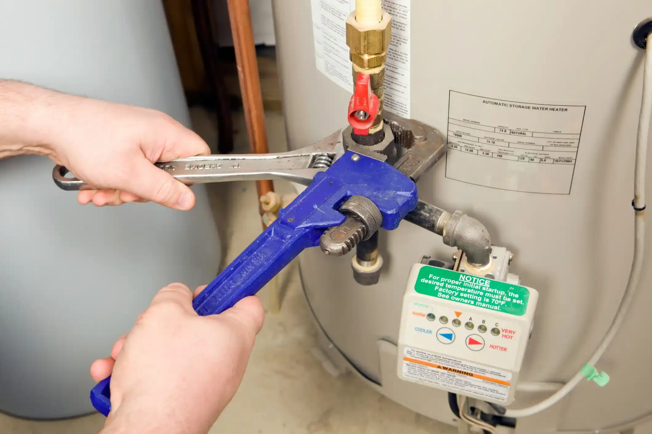  A close-up shot of a person's hands using a large blue pipe wrench and a smaller adjustable wrench to work on the copper piping and valves connected to the top of a water heater. A green and white control box is visible with a "NOTICE" label.