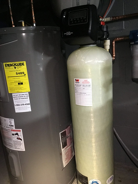  A water heater and water softener are shown side-by-side in a utility room. The gray water heater has a yellow EnergyGuide label on it. Next to it is a tall, light-colored water softener tank with a black digital control head on top. A Warner brand sticker is visible on the water softener tank.