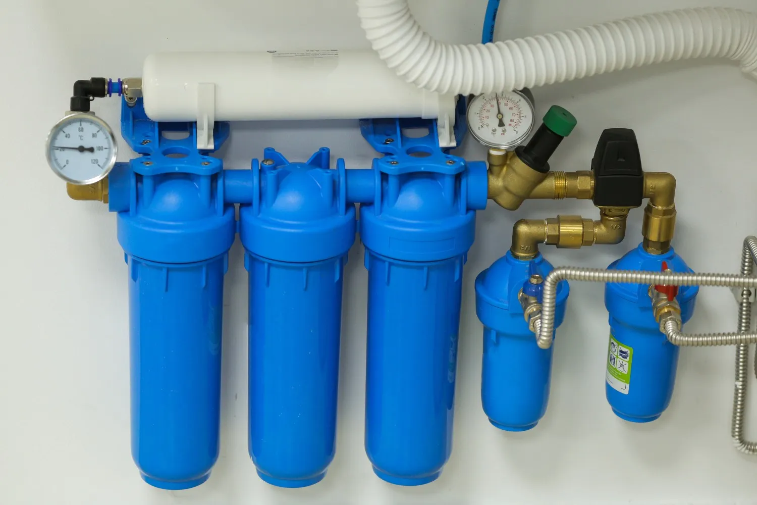A comprehensive whole-home water filtration system with multiple blue filter housings, pressure gauges, and pipes is mounted on a white wall.