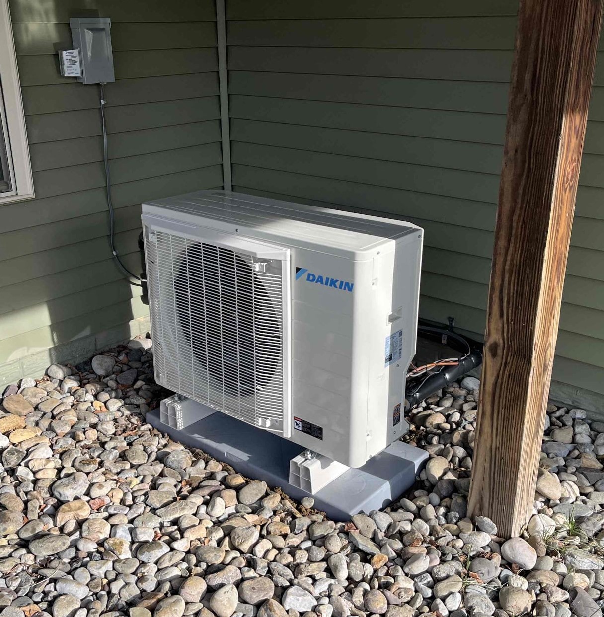  A white Daikin HVAC outdoor unit is sitting on a gray concrete pad on a bed of river rocks next to a green-sided building. A wooden post and some electrical conduits are visible nearby.