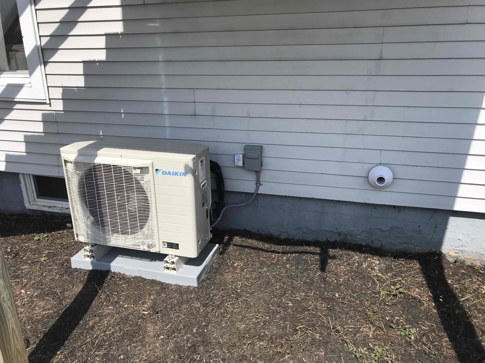  A Daikin outdoor heat pump unit is installed on a gray concrete pad next to a house with light-colored siding. The unit is surrounded by bare dirt and grass. A small electrical conduit box and an exhaust vent are visible on the wall of the house.
