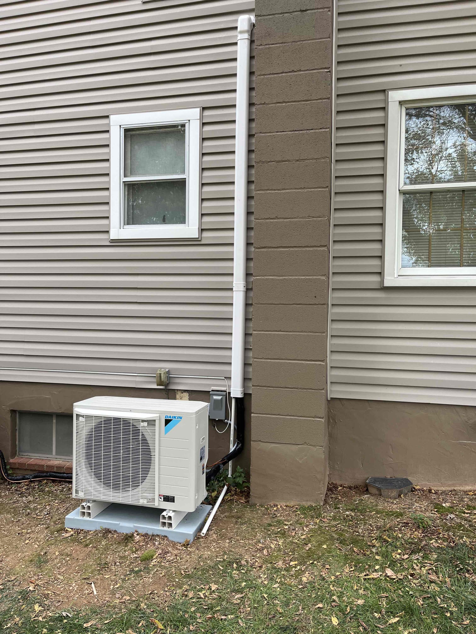  A beige Daikin heat pump is installed on a concrete pad on a patch of grass and dirt. A large white pipe and electrical conduit run up the side of the house next to a painted stone column. There are two windows visible on the beige-sided house.
