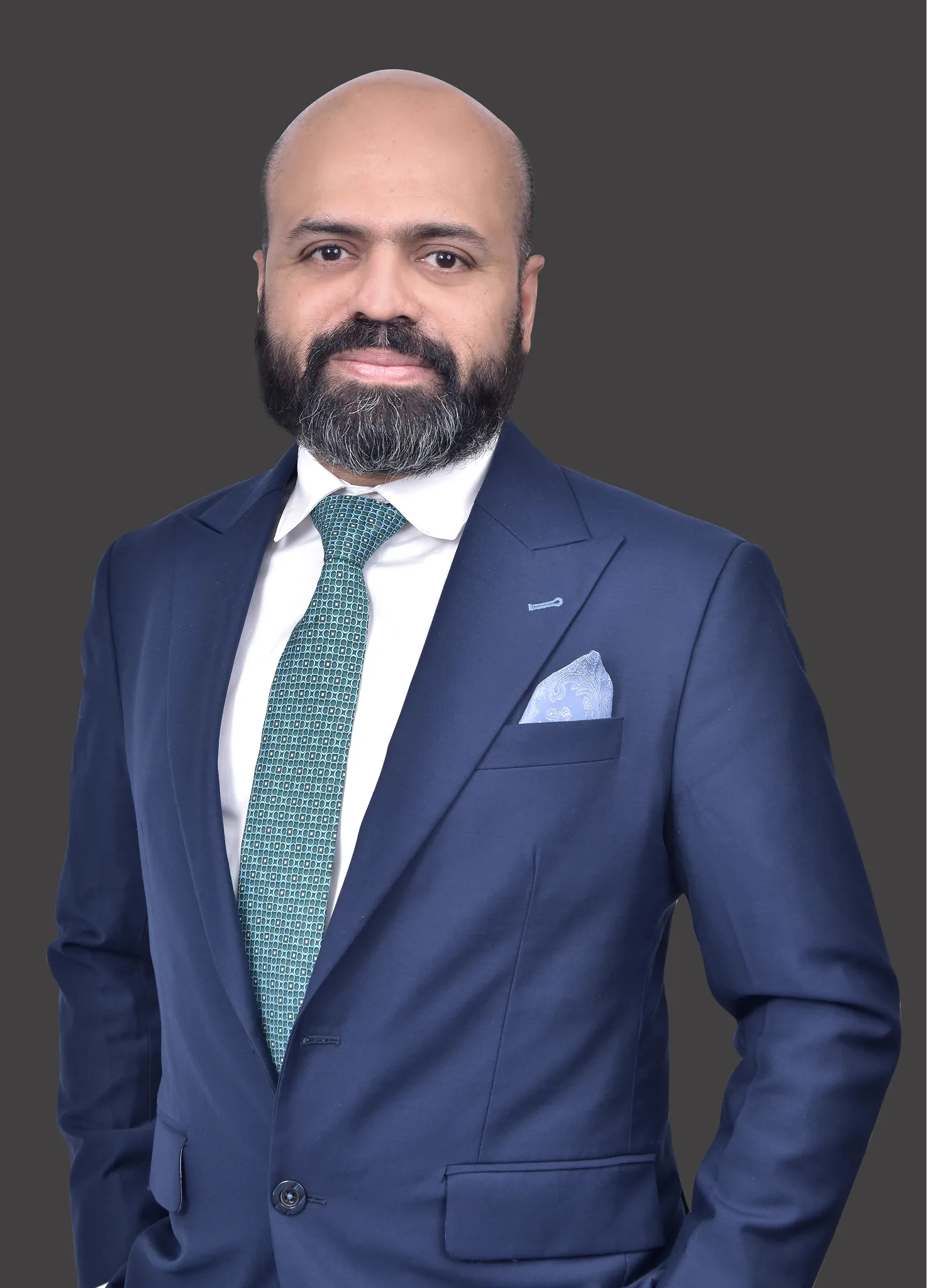 Portrait of a bald man with a beard wearing a navy blue suit, white shirt, teal patterned tie, and a light blue pocket square against a gray background.