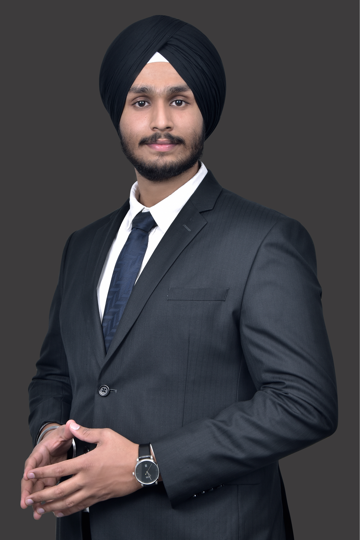 Young man wearing a black turban, black suit, white shirt, and blue tie, posing with hands clasped.