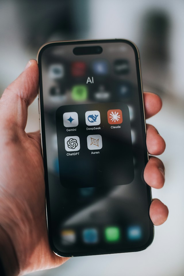 Hand holding a phone showing an AI apps folder with Claude, ChatGPT, Gemini, DeepSeek and Auren
