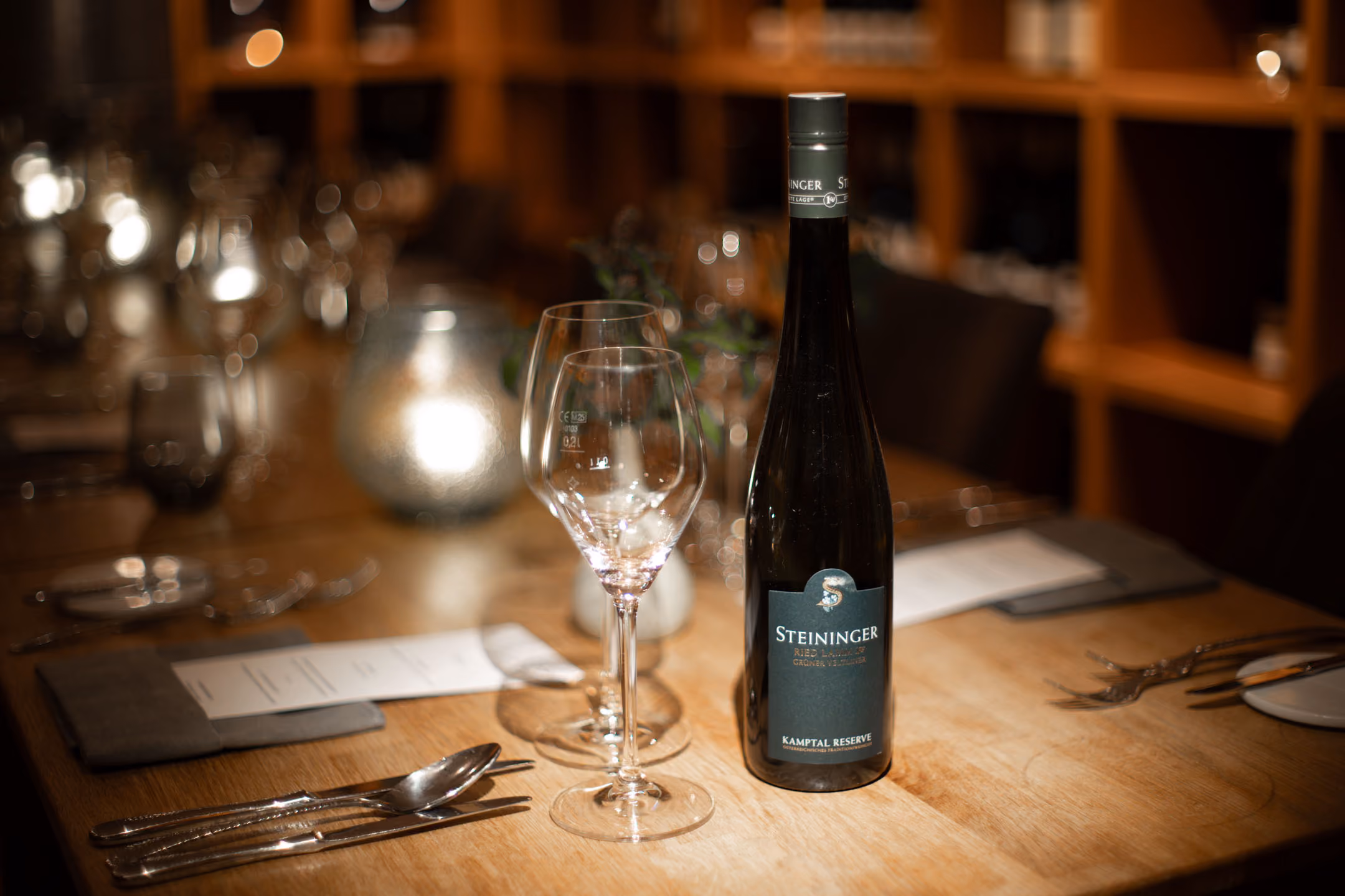 Eine dunkle Weinflasche mit der Aufschrift "Steininger Kamptal Reserve" steht auf einem gedeckten Holztisch neben zwei leeren Weingläsern, Besteck und Menükarten in einem gedämpft beleuchteten Restaurant.