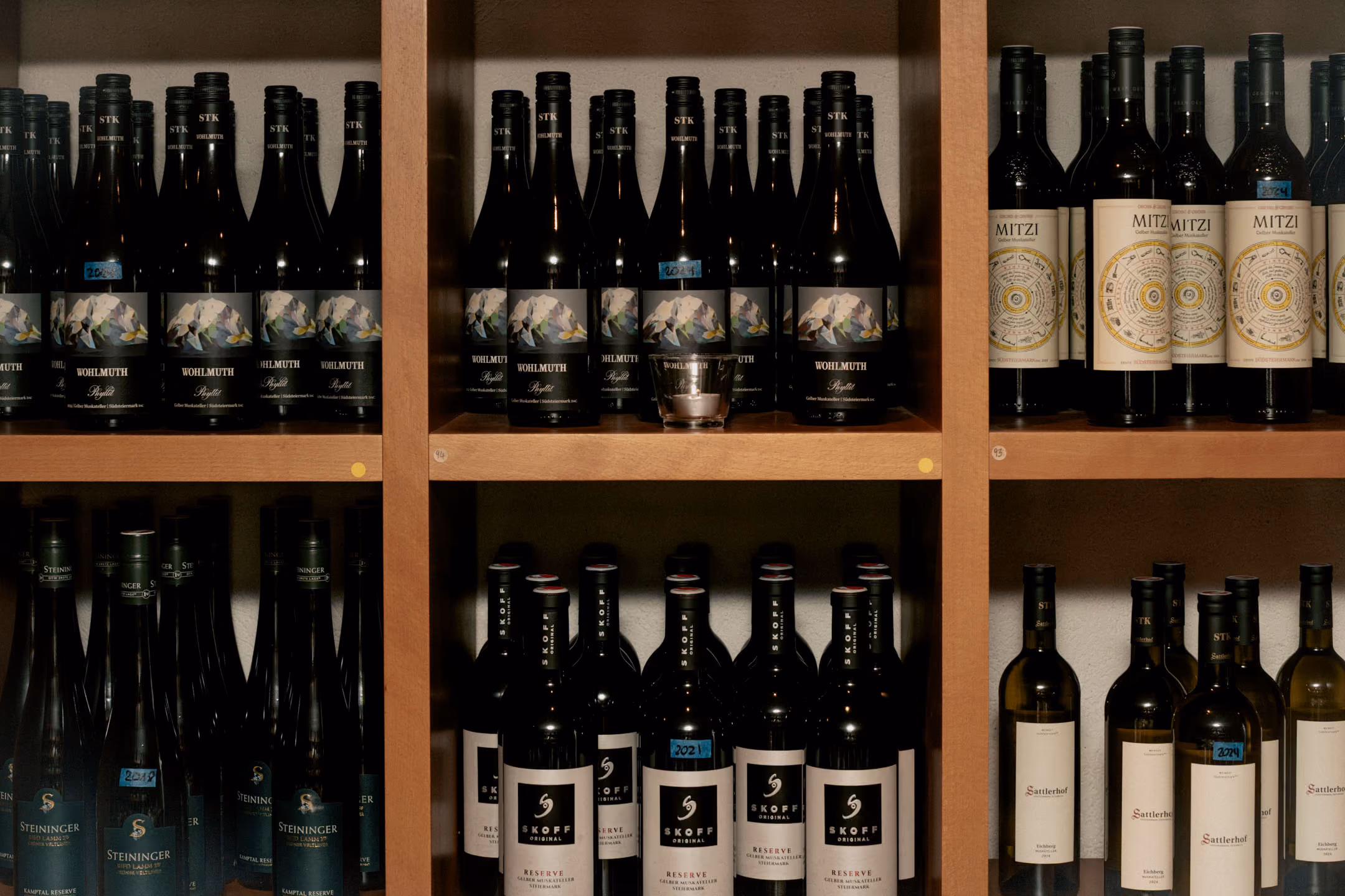 Regal mit drei Reihen verschiedener Weinflaschen, darunter Wohlmuth, Mitzi und Skoff Reserve, angeordnet in einem Holzmöbel.