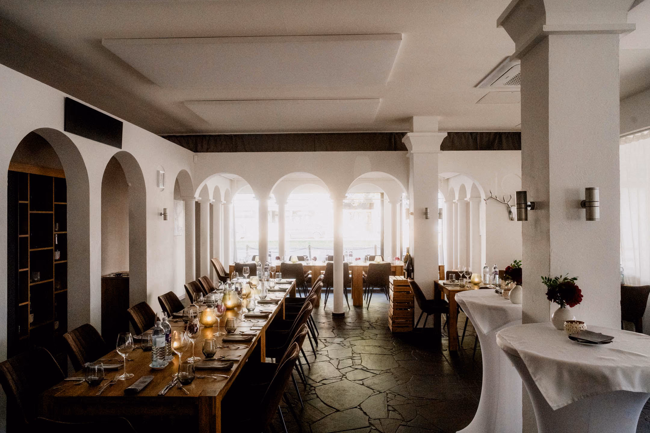Helles Ambiente mit weißen Säulen und Bögen, dunklem Steinfussboden, langen Holztischen und mit weißen Tischtüchern bedeckten Stehtischen, bestückt mit Gläsern, Tellern und Blumenvasen. Dekoriert für eine Hochzeitsgesellschaft neben dem Standesamt Düsseldorf.