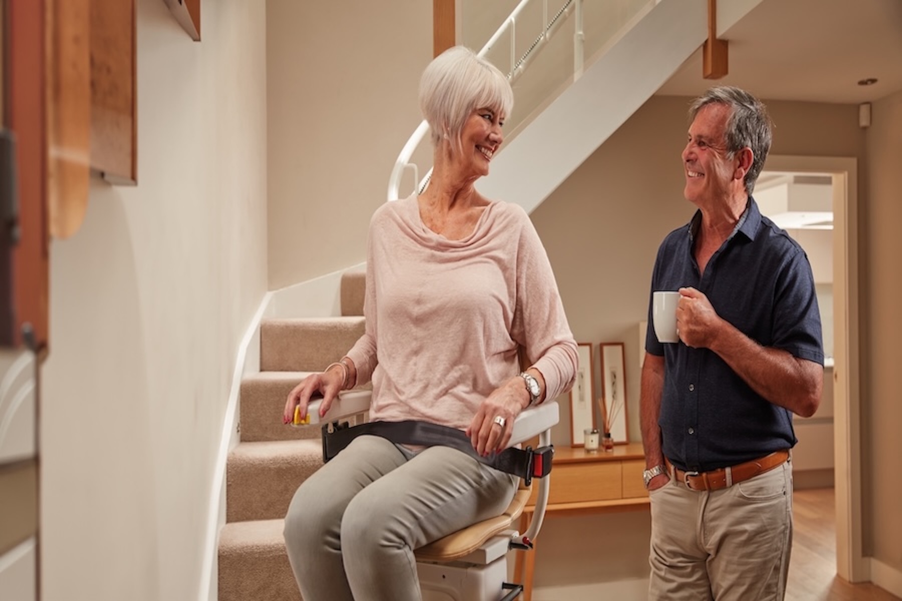 Smiling elderly woman using a stairlift at home while talking to her partner in a modern hallway.