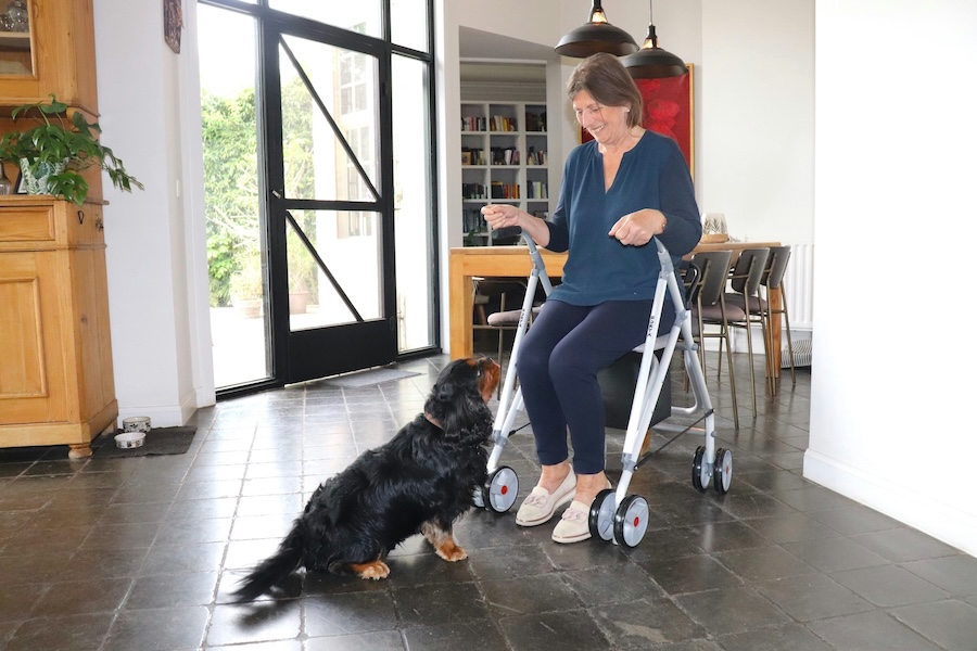 “Smiling elderly woman using a mobility walker indoors while her dog sits beside her.”