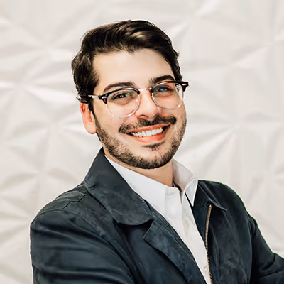 Smiling man with glasses, dark hair, and beard wearing a dark jacket and white shirt against a textured white background.