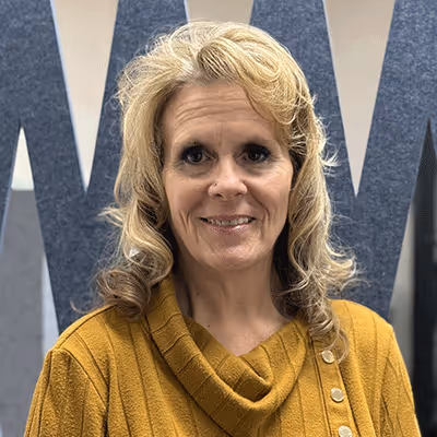 Smiling woman with blonde curly hair wearing a mustard yellow sweater standing in front of a blue and gray wall.