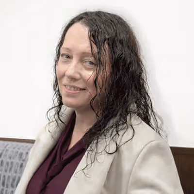 Woman with long curly dark hair wearing a beige blazer and maroon top, smiling slightly against a plain white background.