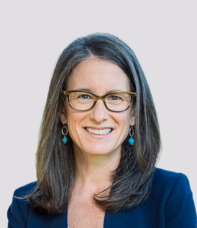 Smiling woman with shoulder-length gray hair, wearing glasses, turquoise earrings, and a dark blazer.