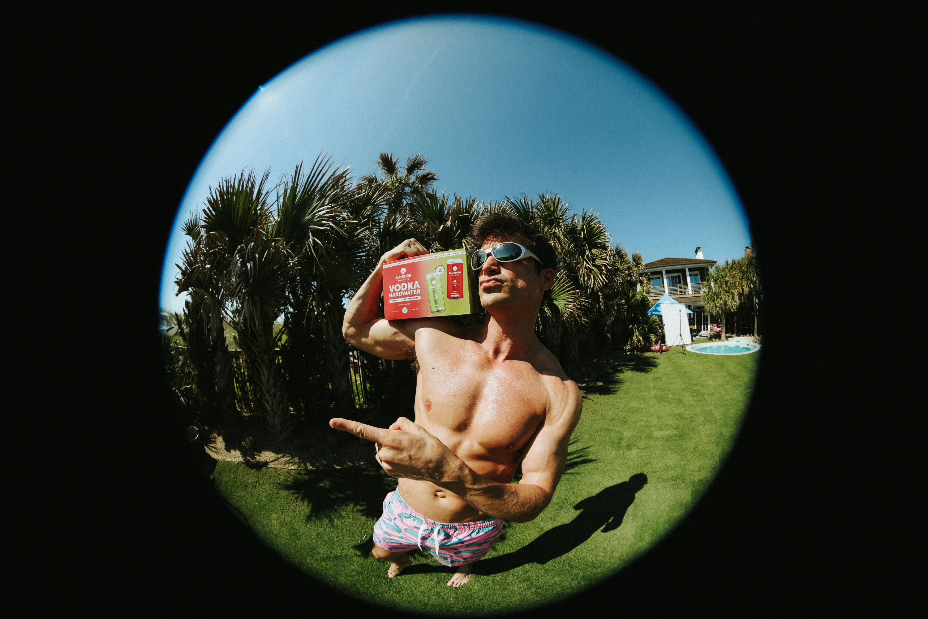 Shirtless man in sunglasses holds vodka pack on shoulder and points in sunny backyard with palm trees.